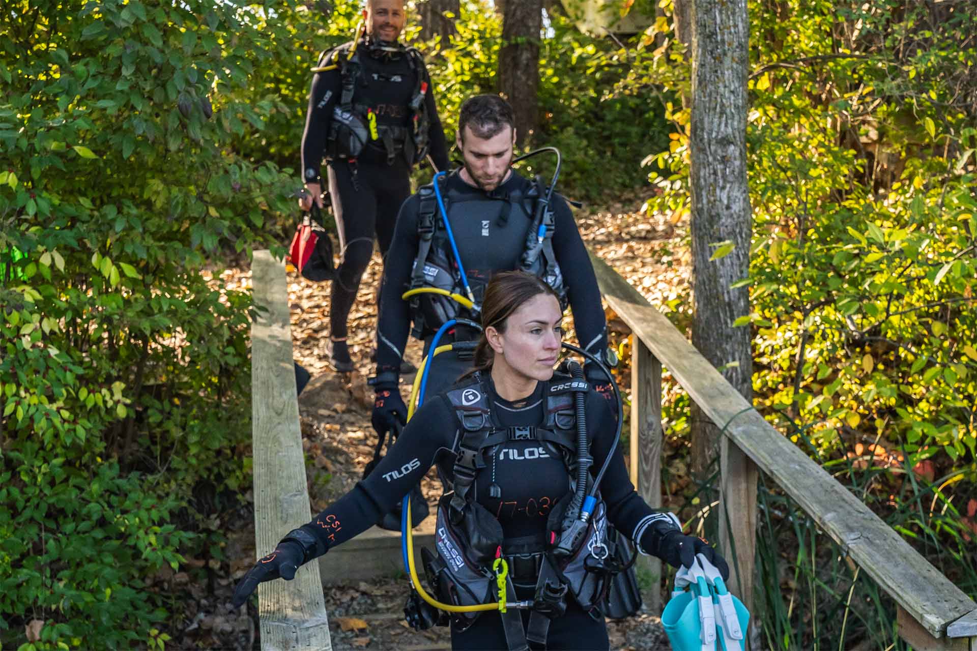 Three scuba divers in full gear and wetsuits walking down a wooded trail toward a dive site — Columbus Scuba PADI Divemaster certification program